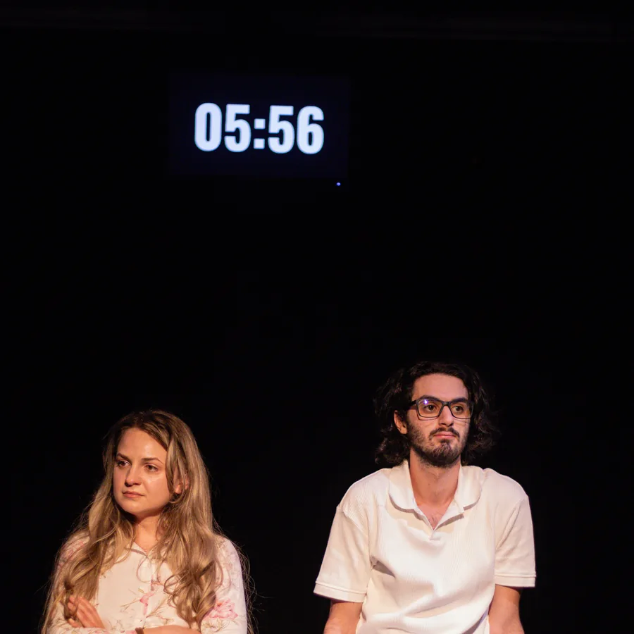 Doi actori pe scenă, cu un cronometru afișat, în spectacolul Au mai rămas 12 minute